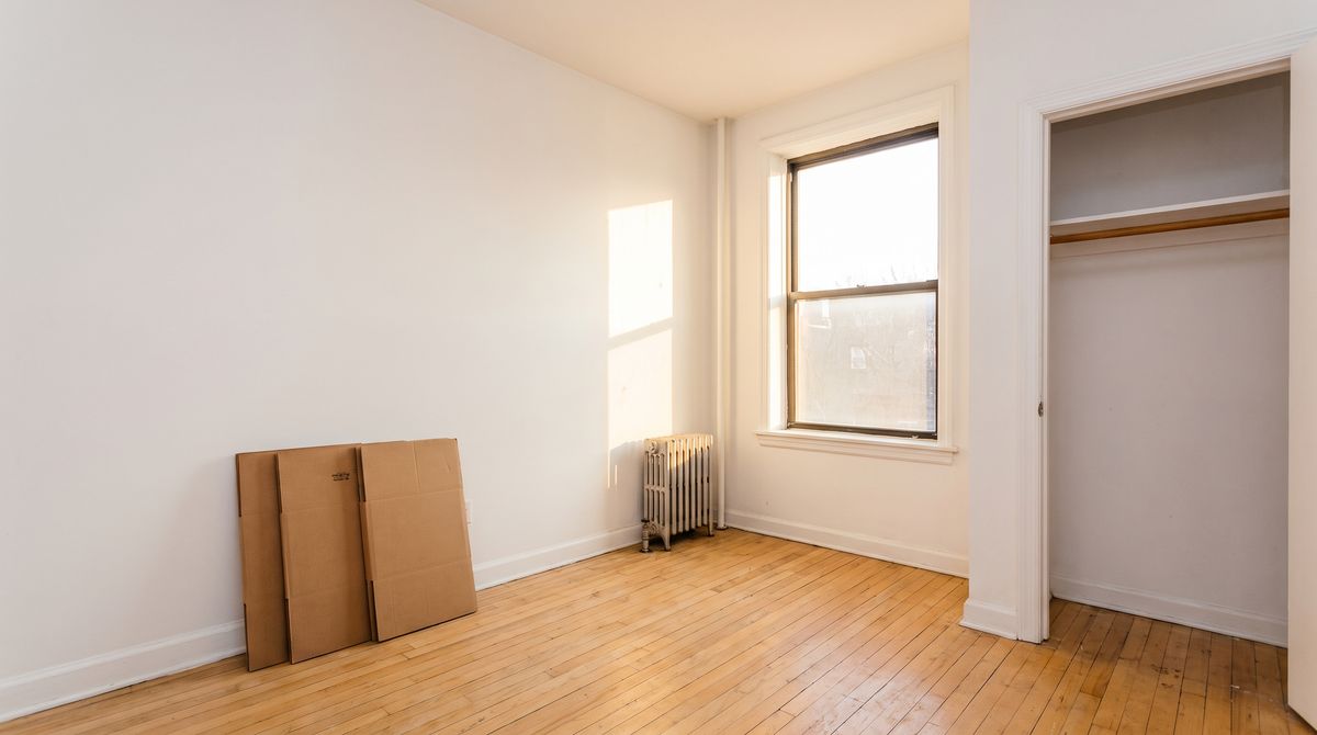 Empty first apartment bedroom on move-in day with bare walls, hardwood floor, a window, an open closet, and flattened moving boxes