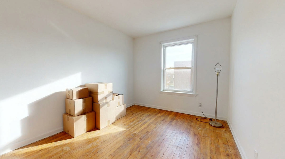 An empty first apartment bedroom on moving day with hardwood floors, a stack of cardboard boxes, and a floor lamp