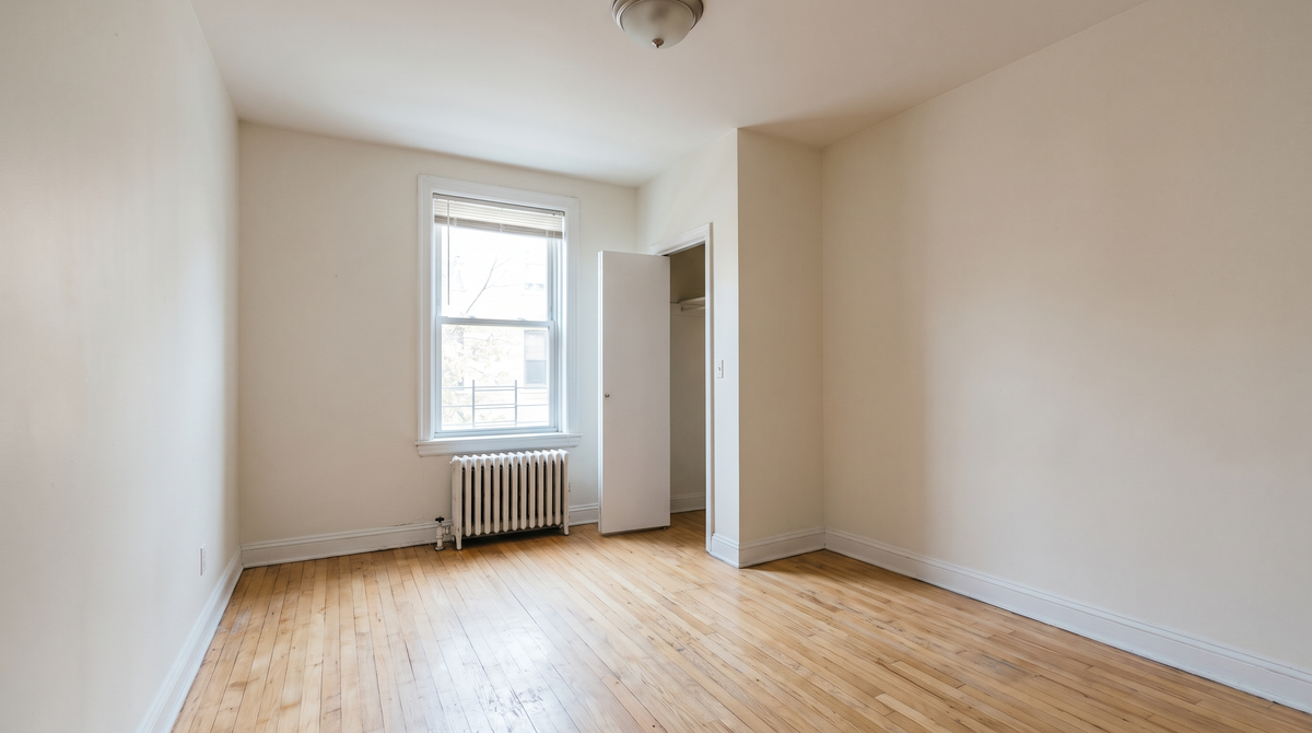 An empty, unfurnished apartment bedroom with hardwood floors, a window with blinds, a radiator and an open closet door