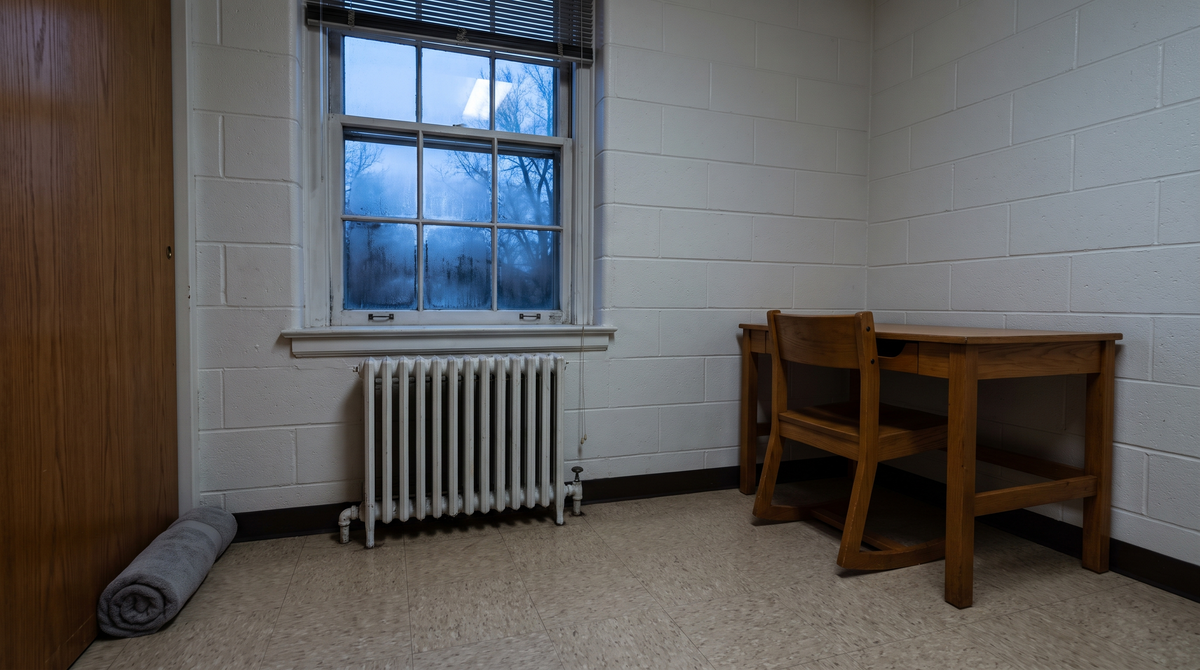 Empty college dorm room corner on a cold evening with a fogged single-pane window, a cast-iron radiator underneath, and a rolled towel blocking the draft under the door